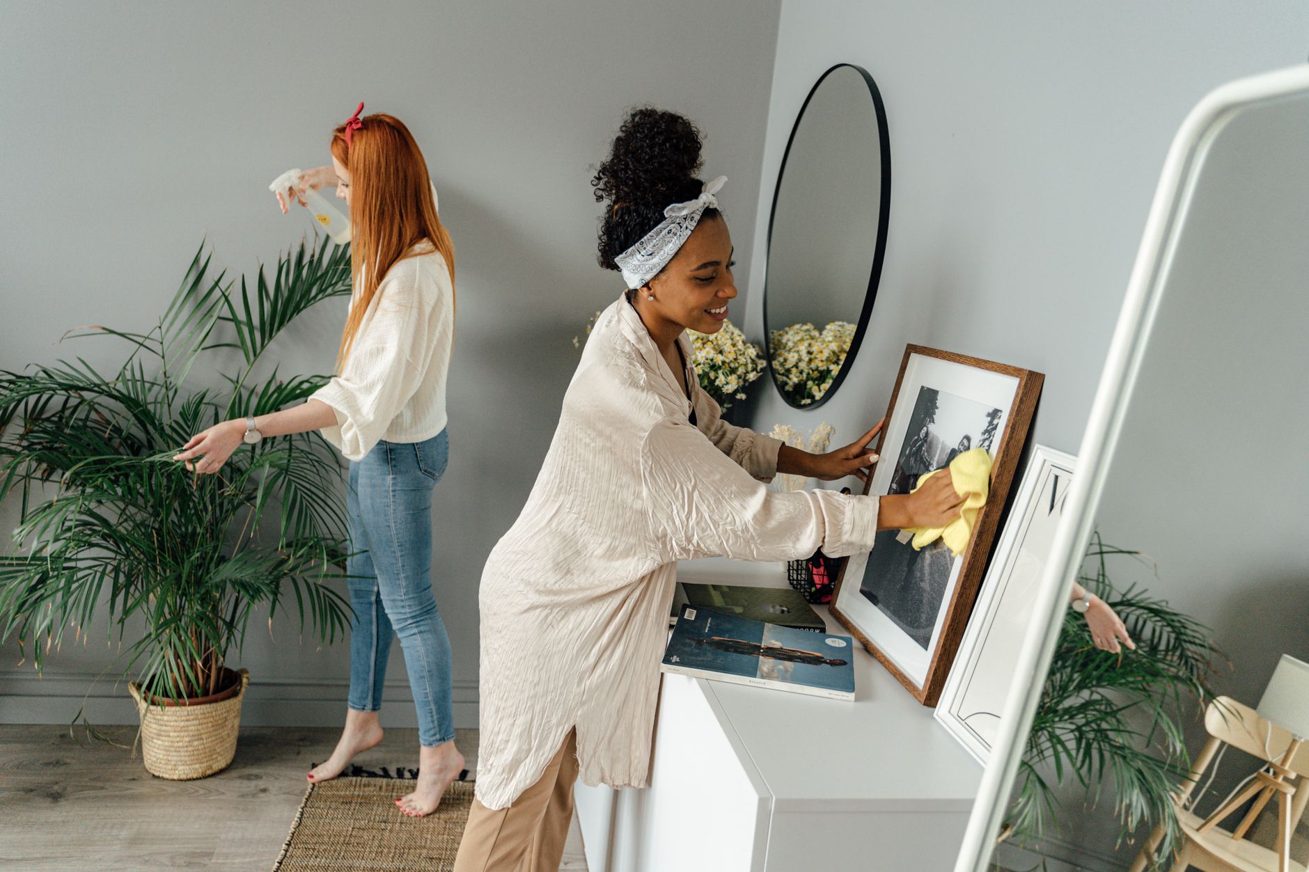 Two women joyfully clean a home interior with plants and decor.